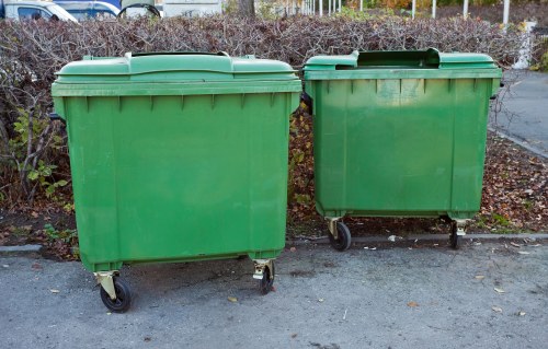 Two large green plastic wheeled rubbish bins are positioned side by side on a paved surface, with the left bin slightly larger and taller than the right one. Both bins are made of durable plastic with a smooth finish and horizontal ridges around their bodies for reinforcement. They feature matching flat, slightly domed lids, which are closed, and are equipped with four black wheels at the base, allowing for easy movement. The bins are situated outdoors on a grey concrete or asphalt surface, with a backdrop of low, leafless bushes and a chain-link fence, indicating a late autumn or winter season in a residential or urban area. The environment appears calm and tidy, consistent with a typical rubbish disposal or garden clearance setting, aligning with services offered by Garden Clearance Sydenham for local rubbish removal in the area near Sydenham. The natural lighting suggests an overcast sky, providing even illumination across the scene without harsh shadows.