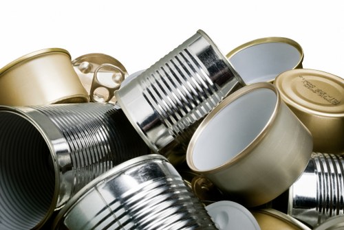 A collection of metal and tin cans stacked together, including both opened and unopened examples, with some cans showing ridged sides and smooth finishes. The cans vary in colour, primarily silver, gold, and metallic hues, with some lids visible on the open cans. They are arranged on a surface, possibly in an outdoor or indoor setting such as a garden or a storage area, with no additional objects or background details visible. Lighting highlights the reflective quality of the cans' surfaces, contributing to a clear and detailed view of their textures and finishes, consistent with waste removal or recycling context in Sydenham. Garden Clearance Sydenham handles such rubbish, including metal and tin containers, for proper disposal or recycling.