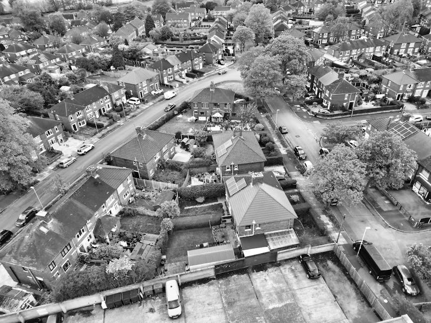 An aerial view of a residential neighbourhood showing a variety of houses, each with distinct features and different roofing materials, including tiled and metal roofs in shades of grey. The scene cap